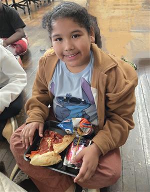  child sitting on the floor smiling with a plate of food in lap